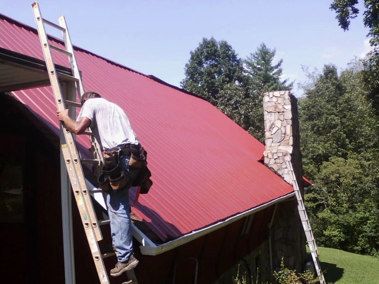 Roofer on a ladder beside a red metal roof and stone chimney — Soffit & Fascia Repair by Roofing Brothers in Colorado Springs